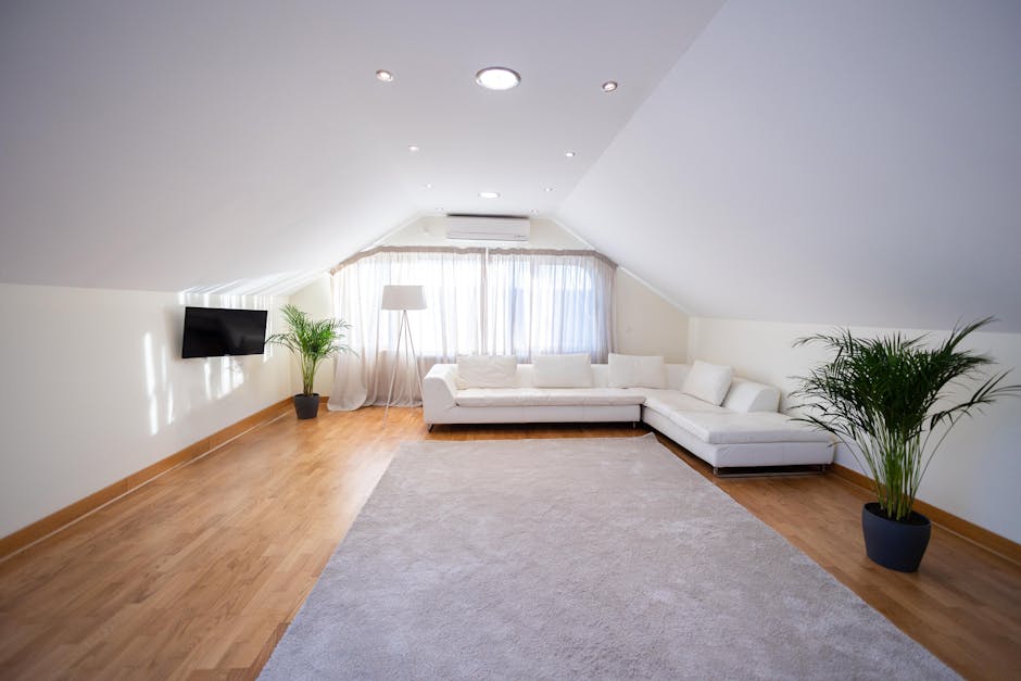 A spacious living room featuring light wooden flooring and a cream-colored sectional sofa positioned along the windows, which are adorned with sheer white curtains allowing natural light to fill the s