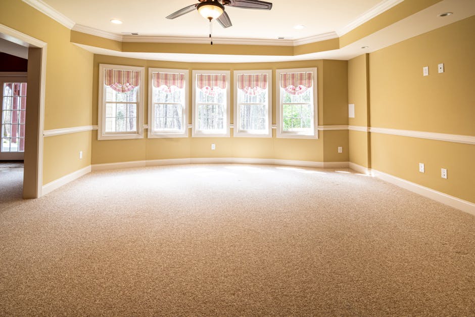 A spacious living room featuring light wooden flooring and a cream-colored sectional sofa positioned along the windows, which are adorned with sheer white curtains allowing natural light to fill the s